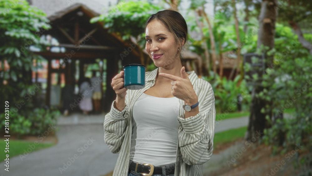 Fototapeta premium Woman holding teal mug and pointing at camera at building entrance, wearing white tank and striped shirt; confidence.