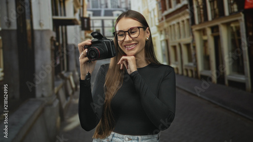 Obraz na płótnie Woman holding dslr camera and smiling while raising the camera on a cobblestone street; happy creativity