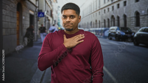 Konstfotografi Hispanic man places hand on chest on a street wearing a maroon shirt and beard visible; sincerity humility