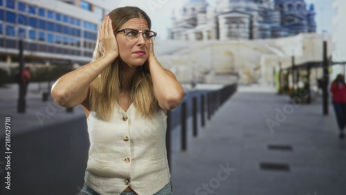 Cuadro en lienzo Woman covers ears with hands and glasses on street near monument and bollards, wearing sleeveless vest and jeans; stress discomfort