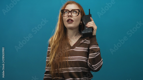 Foto Redhead woman holds retro cellphone to ear in studio while pouting, glasses and striped sweater visible; annoyance commentary