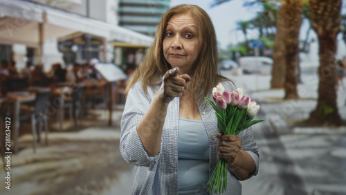 Canvastavla Senior woman holding a bouquet of pink tulips points a finger toward camera at a street cafe; gentle reproach