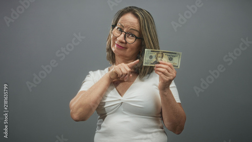 Cuadro en lienzo Hispanic senior woman with grey hair and glasses holding a twenty dollar banknote and pointing finger to bill in studio, wearing white top; playful savings