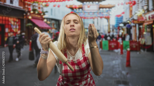 Canvastavla Woman chef in red check apron holds rolling pin and makes pinched finger gesture in a busy street market; annoyance challenge