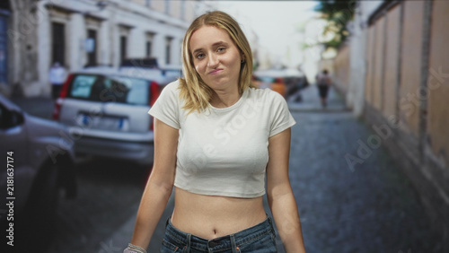 Картината върху платно Woman bare stomach in white crop t shirt shrugs shoulders on a cobblestone street beside parked cars and buildings; uncertainty
