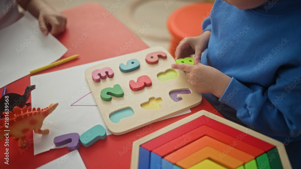 Fototapeta premium Boy placing wooden number puzzle pieces with hands on red table in preschool, colorful rainbow stack, toy dinosaurs, pencil and drawing paper visible; concentration learning playtime.