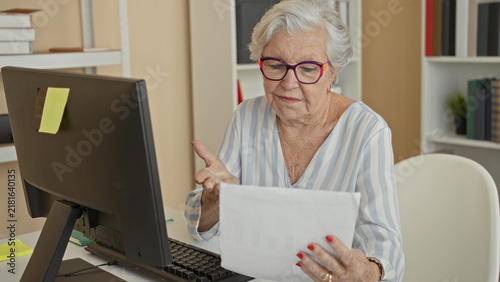 Foto Older woman holding paper and reading at a computer desk in a building with hand raised and hand to chin gesture; concern focus