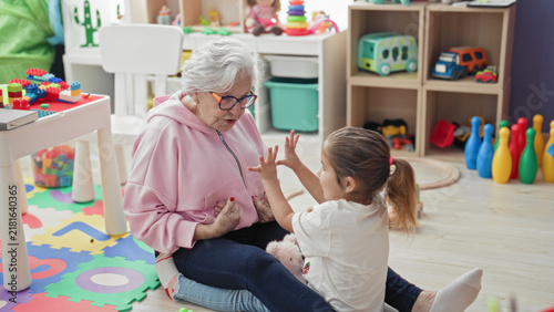 Cuadro en lienzo Elderly woman reaches arms to hug little girl on a toy strewn playmat inside a building; affection bonding