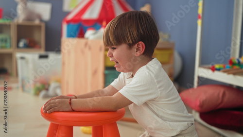 Konstfotografi Boy crying with tears on cheek and open mouth, hands gripping an orange stool amid toys and a small tent inside a playroom building; sadness