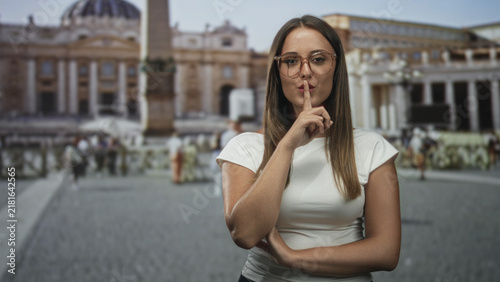 Картината върху платно Young woman finger to lips shushing in a cathedral square near a historic basilica; mystery quiet travel intrigue