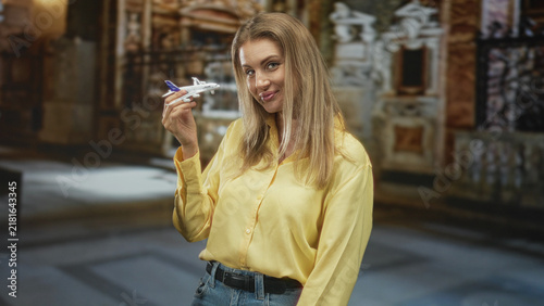 Принти на полотні Blonde woman holding a toy airplane in her hand, smiling and posing in an ornate historic building interior with decorative walls and marble floor; travel optimism