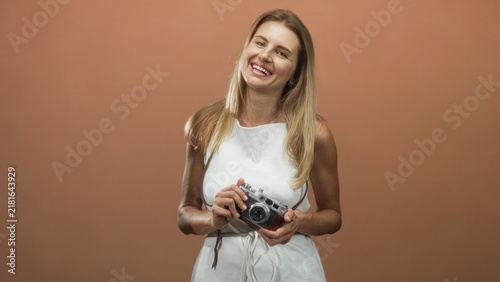 Foto Blonde woman holding vintage rangefinder camera with bare hands, smiling and tilting head while wearing white dress and camera strap visible in studio; joy