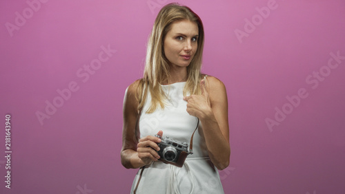 Bild auf Leinwand Blonde woman holding vintage camera in white sleeveless dress, thumb pointing to chest, leather strap visible, studio pink backdrop; confidence travel