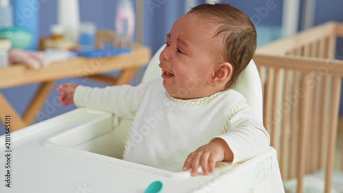 Принти на полотні Hispanic baby boy crying in highchair with food on mouth and arm outstretched in a building nursery; feeding distress hunger
