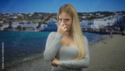 Картината върху платно Woman adjusts glasses with fingertip and crossed arms near seaside building and pebble shore; thoughtful doubt