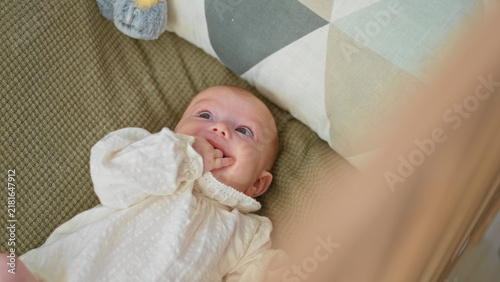 Girl lying in crib with fingers in mouth wearing white gown, looking up toward mobile and plush toy near patterned pillow and blanket; gentle comfort.