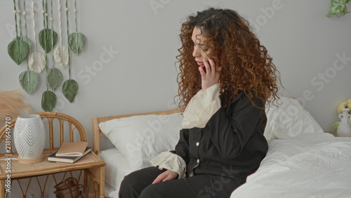 Фотография Woman sitting on bed touching her face and tousling curly hair beside a white duvet in a building; quiet contemplation pause