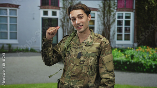 Man in camouflage uniform wearing dogtags, bare thumb pointing over shoulder in a casual salute gesture toward a building entrance, smiling; pride duty.