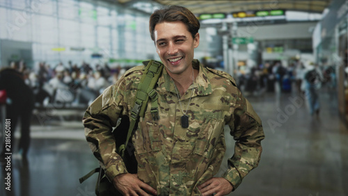 Foto Man soldier in camouflage uniform carrying backpack and dog tags with hands on hips in busy airport concourse; pride homecoming