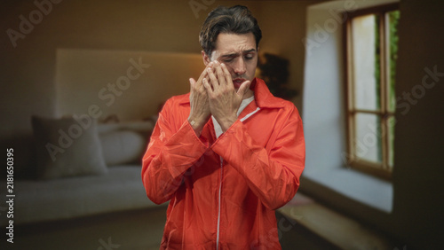 Photography Man in orange inmate jumpsuit inspects palms and rubs eye while standing by a window in a building; remorse reflection