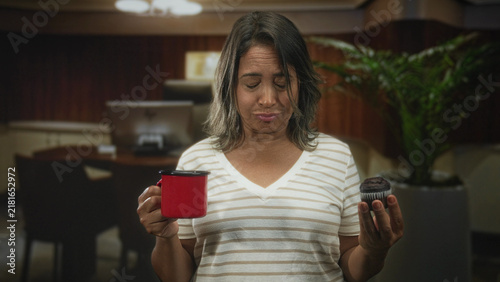Foto Woman holding red cup and chocolate muffin with eyes closed and grimace in building; hesitation disgust