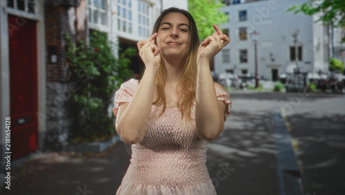 Foto Woman with fingers crossed and eyes closed, wearing a pink smocked top, standing in a leafy urban street with buildings and bicycles; hopeful anticipation