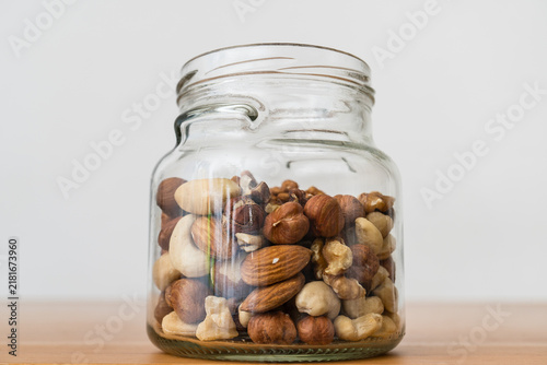 Glass jar filled with almonds, walnuts, cashews and hazelnuts on wooden table against light background.