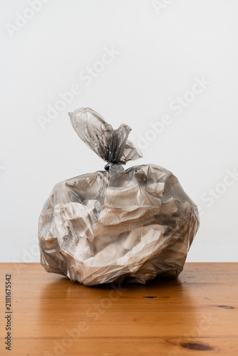 Close-up photo of tied transparent plastic bag filled with household trash on wooden table. Crumpled packaging and waste are visible through bag.