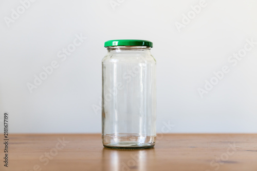 Front view of empty clear glass jar with green metal lid on wooden table against minimalist light grey background with copy space.