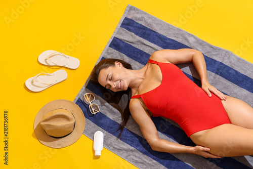 Cuadro en lienzo Smiling tanned woman in swimsuit sunbathing on towel and beach accessories on ye