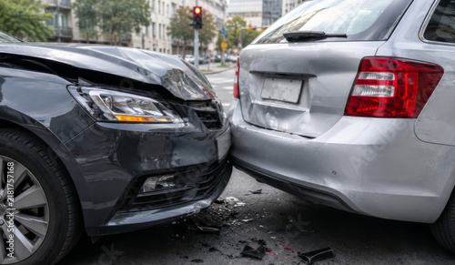 Two cars with damaged bumper after traffic accident on city street. Black car crashed into silver car rear at intersection with traffic light. Auto collision, insurance claim and road safety concept.