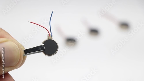 Coin type micro vibration motor with red and blue wires held by tweezers with other motors blurred in background