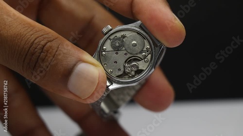 Mechanical watch movement with damaged spring held in fingers showing exposed gears and internal parts needing repair