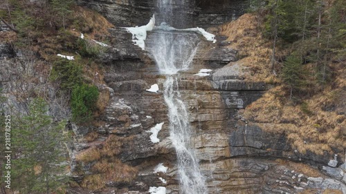 Waterfall cascades down rocky cliff face. Spray mist rises where water meets mossy stones. Tall pines cling to mountain sides