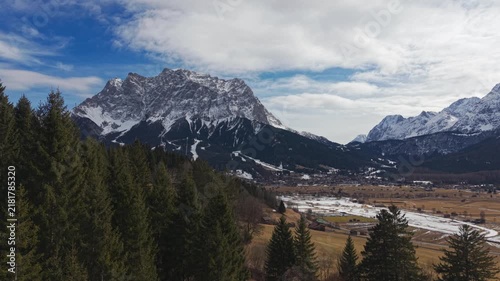 Snow-capped peaks rise above green pines and golden meadows. Vast clouds drift across a bright blue sky