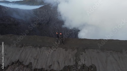 Aerial view of two hikers walking along the narrow edge of an active volcano crater with smoke rising