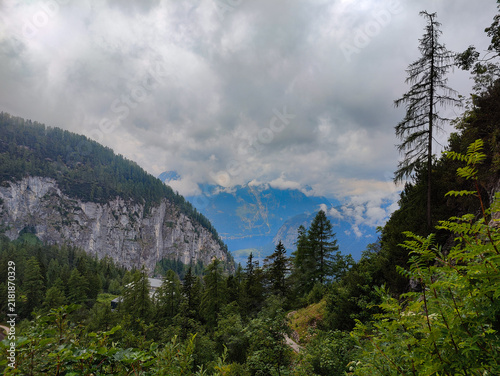 A serene mountain valley shrouded in clouds with lush forests, rocky cliffs, and distant snow-capped peaks. Vibrant foliage creates a tranquil alpine landscape perfect for nature enthusiasts.