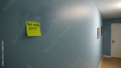 Yellow sticky note with 'Get ready 2027' message on a blue wall in a hallway with soft ambient light, created with generative ai