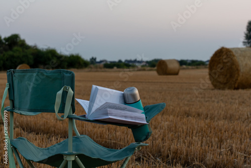 Bild auf Leinwand Open book and reusable thermos placed on a portable chair in a harvested field with hay bales