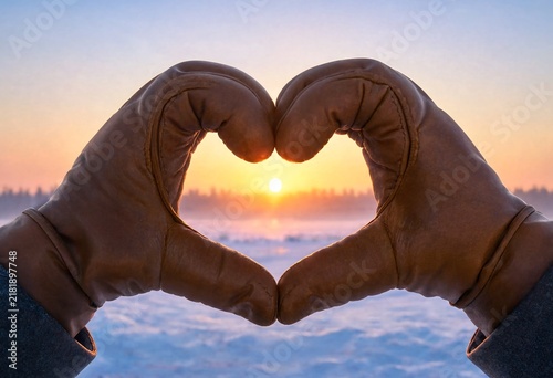 Hands forming heart shape against winter sunrise with soft golden light and mist