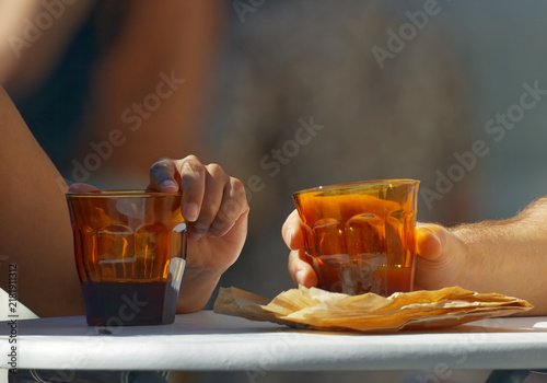 Two hands holding glasses of amber coffee backlit by warm sunlight, a crumpled kraft paper wrapper resting between them at the small, community-minded Hermanak farmers market in Prague.
