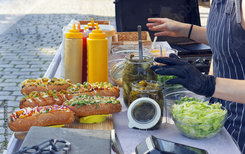 A gloved vendor prepares gourmet hot dogs beside condiment bottles, pickle jars and fresh lettuce on a cobblestone street at the Naplavka market in Prague.
