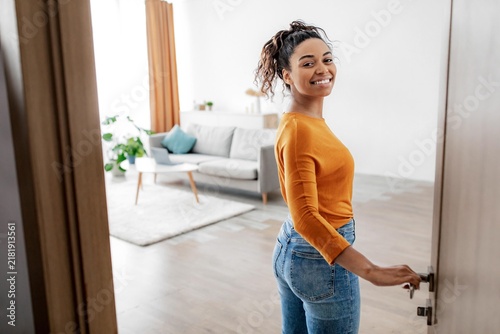 Joyful Smiling Black Woman Opening Door With Happiness, Standing In Modern Living Room, Welcoming Guests, In Cozy Home Environment. Home Interior Concept.