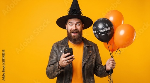 Man with witch hat holding black and orange air balloons for halloween party surprised and sending a message