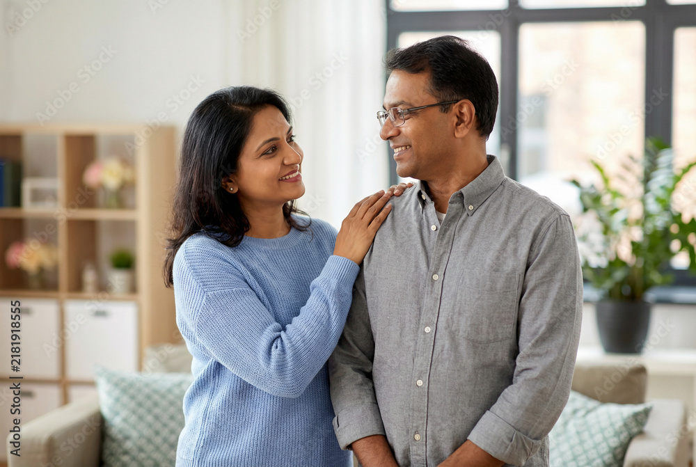 Obraz premium Happy Indian couple smiling at each other standing in modern living room with bright natural light at home