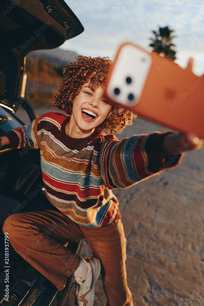 Obraz premium Woman selfie smile curly hair outdoors car lifestyle, young adult cheerful expression, striped sweater, leaning near open vehicle door, warm sunlight, urban summer travel relaxed mood
