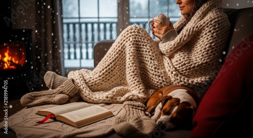 Woman wrapped in a cozy knitted blanket holds a warm mug while sitting near a fireplace with her dog.