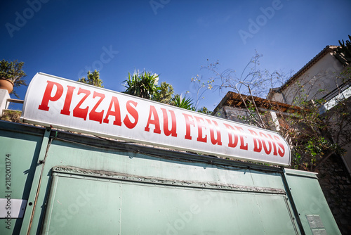 A large sign reading PIZZAS AU FEU DE BOIS, meaning wood fired pizza in french advertises above a roadside food stand, on a common French takeaway and pizzeria offering under a clear blue sky.