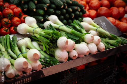 Close-up of bunches of young onions, also known as green onions or scallions, displayed for sale on a market stall, with tomatoes and cucumbers visible in the background.