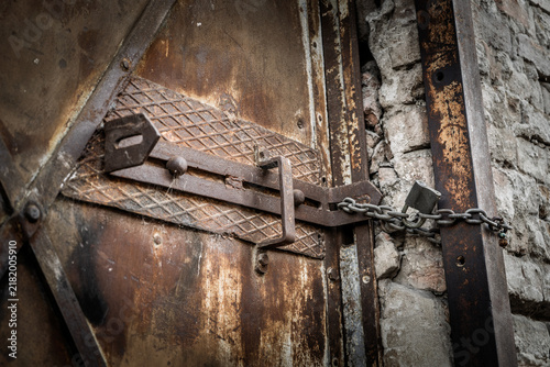 фотографія Close-up of a heavy rusty metal latch secured with a chain and padlock on an old weathered door, showing corrosion, aged hardware and rough masonry around a locked and deteriorating entrance